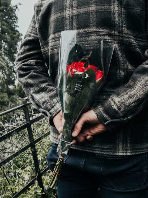 A person wearing a dark plaid jacket holding a bouquet of fresh red and white roses wrapped in clear plastic, with dark green leaves visible among the flowers. The bouquet is arranged neatly with the 