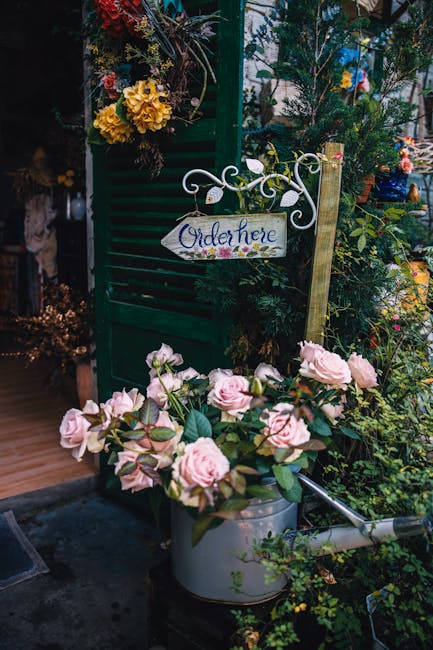 A lush floral display featuring a bouquet of soft pink roses with velvety petals and fresh green foliage, arranged in a silver watering can placed outdoors amidst abundant greenery and flowering plant