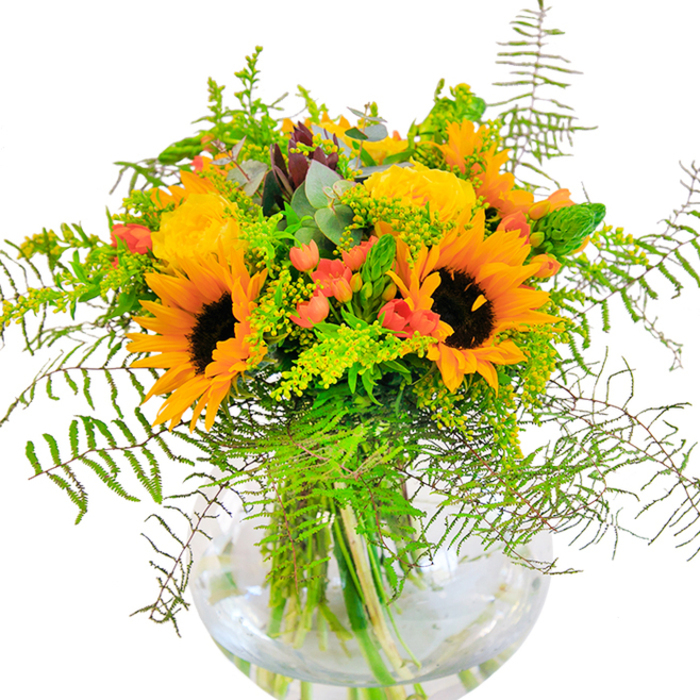 The first impression this bouquet evokes is one of radiant, golden energy-an uplifting spectacle reminiscent of Chadwell Heath's brightest summer days. Sunflowers, their lively yellow-orange petals curling about deep brown centres, stand tall amongst buttery garden roses whose densely packed spirals lend an air of timeless grace. Glossy crimson berries emerge here and there, infusing the composition with playful warmth and contrast, much like the vibrant streetside displays near Chadwell Heath's bustling Broadway. The backdrop of lush, emerald foliage includes soft fern fronds that spill in gentle cascades, echoing the untamed edges of local natural spaces, while slender spikes and soft golden filler blossoms contribute delicate, textural depth. Eucalyptus leaves with their silvery-blue overtones nestle between blooms, adding a hint of rustic charm to the display. The round, crystal-clear vase cradles the submerged stems, highlighting the precise architecture of the arrangement and reflecting light in bright, refreshing dashes across the table. Up close, the bouquet's layered textures combine suede-like sunflower petals, velvety roses, and crisp greens, while a subtle, invigorating fragrance whispers promises of summer fields and new beginnings. Designed as an exuberant gesture for birthdays, milestones, or festive occasions in Chadwell Heath, this arrangement is a heartfelt reminder of happiness shared and moments worth cherishing.